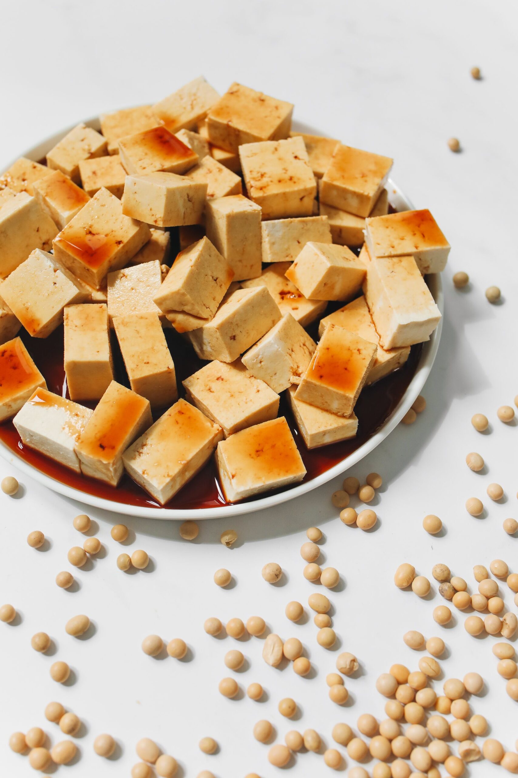 Appetizing tofu cubes drizzled with soy sauce on a white plate, surrounded by raw soybeans.